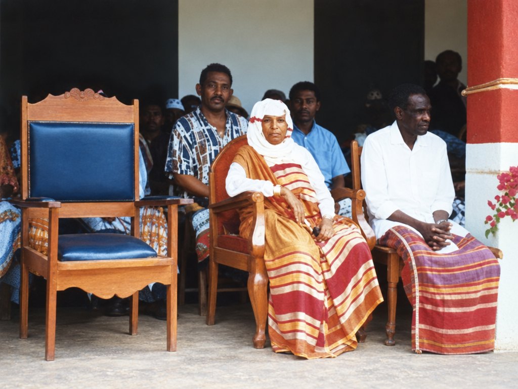 Queen Amina Said seated on a wooden chair in a white head covering and a striped wrap, her husband beside her in a white shirt, with men of Namakia behind them